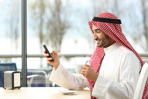 Side view of an arab man texting in a smart phone in a coffee shop with a window with a sunny day in the background